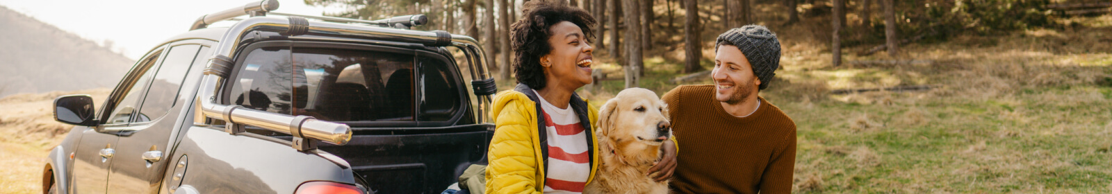 Man, woman, and dog sitting the their truck bed in the wilderness smiling.